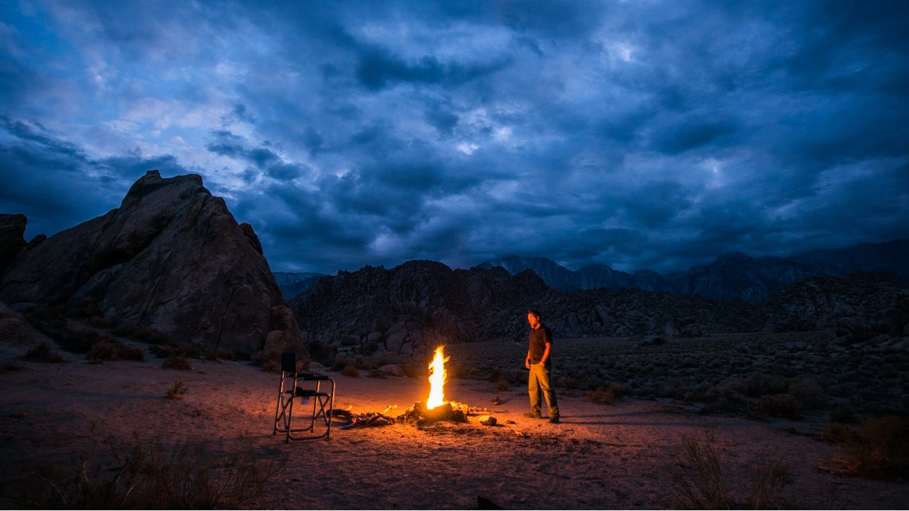 Person by a campfire in a desert landscape under a dramatic blue sky.